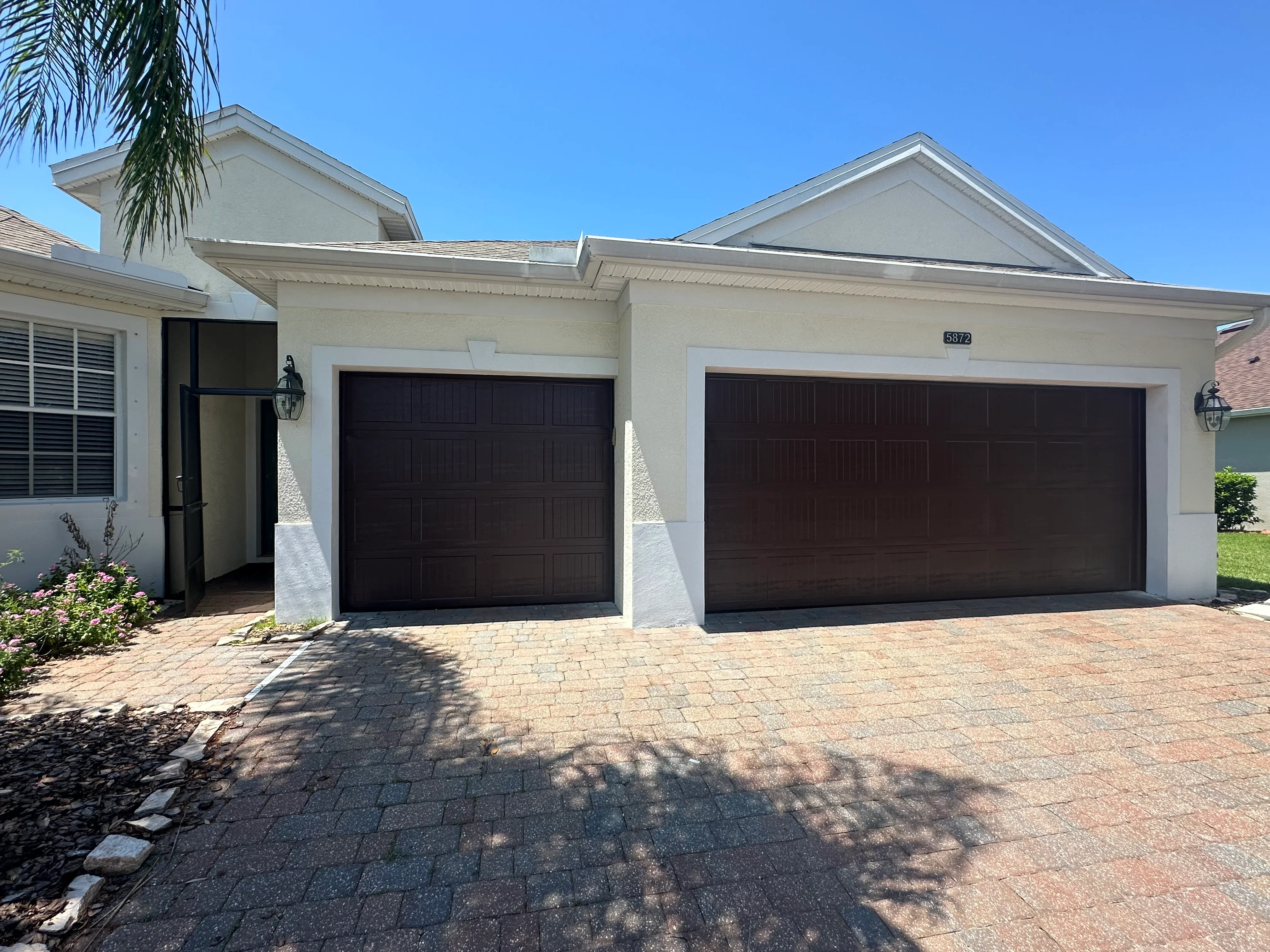 Two dark brown garage doors installed on cream stucco home with white trim and paver driveway